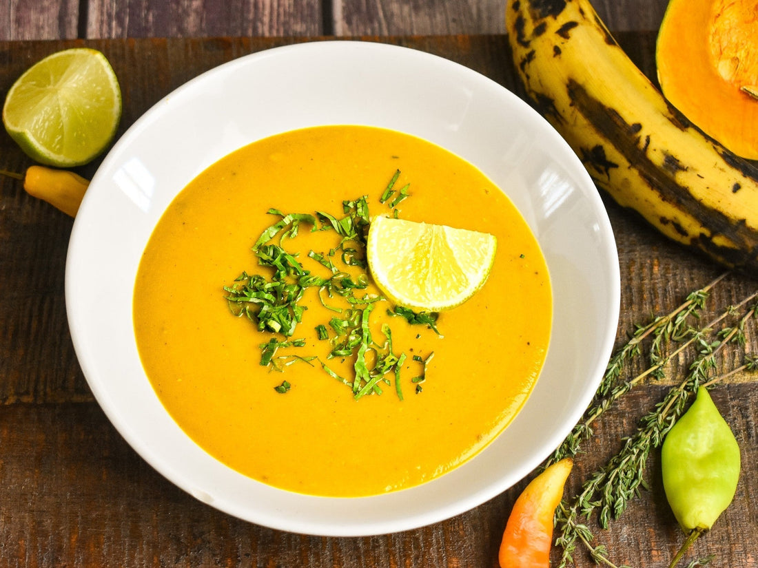 a soothing hormone-friendly bowl of creamy caribbean pumpkin and plantain soup plated in a white bowl on a wooden cutting board surrounded by fresh caribbean ingredients like ripe plantain